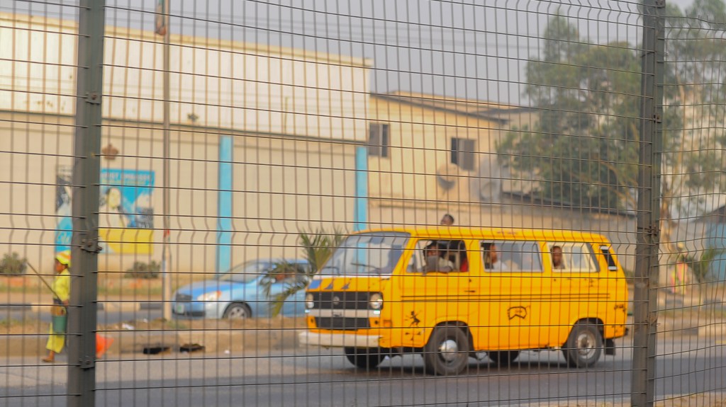 A yellow danfo minibus travelling along a Lagos street, photographed through a roadside fence.